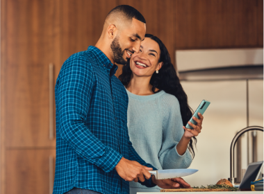 Man cooking while woman smiles and holds her phone.