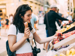 Young tourist woman eating an ice cream and buying some fruit in a street market while walking arround the city. Unrecognizable people at the background