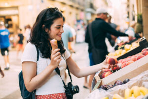 Young tourist woman eating an ice cream and buying some fruit in a street market while walking arround the city. Unrecognizable people at the background