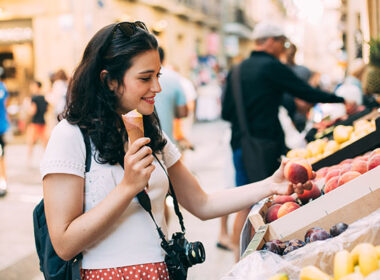 Young tourist woman eating an ice cream and buying some fruit in a street market while walking arround the city. Unrecognizable people at the background