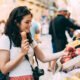 Young tourist woman eating an ice cream and buying some fruit in a street market while walking arround the city. Unrecognizable people at the background