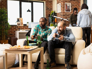 Young diverse men seated on sofa with wireless controllers, playing video games on television with multiethnic group of friends.