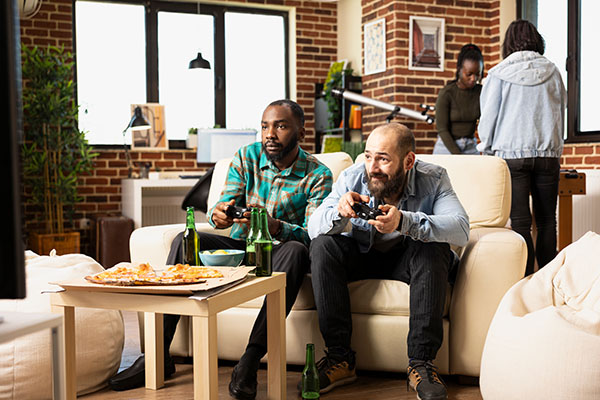 Young diverse men seated on sofa with wireless controllers, playing video games on television with multiethnic group of friends.