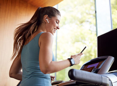 Woman Exercising On Treadmill Wearing Wireless Earphones And Smart Watch Checking Mobile Phone