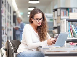 Young female student studying in a college library, sitting at desk with laptop and smartphone