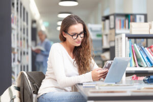 Young female student studying in a college library, sitting at desk with laptop and smartphone