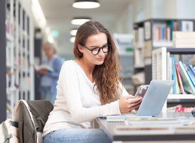 Young female student studying in a college library, sitting at desk with laptop and smartphone
