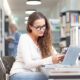 Young female student studying in a college library, sitting at desk with laptop and smartphone