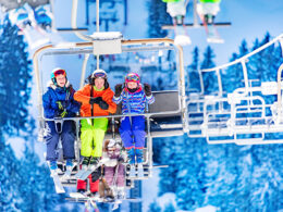 Three girls in colorful outfits sit on ski lift talking and smiling together lifting on top of the mountain