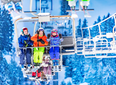 Three girls in colorful outfits sit on ski lift talking and smiling together lifting on top of the mountain