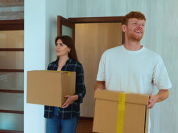 Portrait of Caucasian cheerful young couple in good mood entering their new apartment holding boxes in hands and putting them down on the floor.