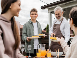 bearded father and young adult son holding plates, mother and teenage sister on foreground, backyard