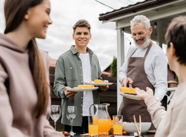 bearded father and young adult son holding plates, mother and teenage sister on foreground, backyard