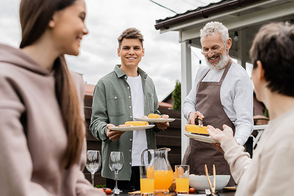 bearded father and young adult son holding plates, mother and teenage sister on foreground, backyard