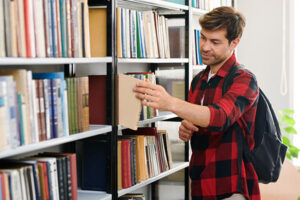 Young student with backpack taking one of books from shelf while visiting college library