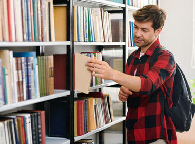 Young student with backpack taking one of books from shelf while visiting college library