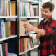 Young student with backpack taking one of books from shelf while visiting college library