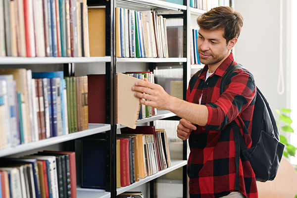 Young student with backpack taking one of books from shelf while visiting college library