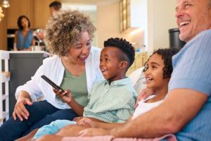 Grandparents And Grandchildren Sitting On Sofa Watching TV At Home With Parents In Background