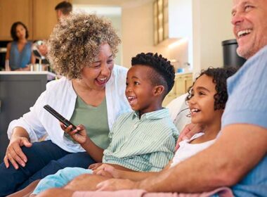 Grandparents And Grandchildren Sitting On Sofa Watching TV At Home With Parents In Background