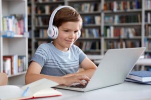 Little boy with headphones reading book using laptop in library