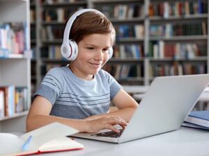 Little boy with headphones reading book using laptop in library
