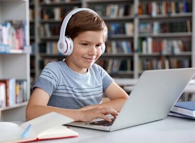Little boy with headphones reading book using laptop in library