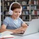 Little boy with headphones reading book using laptop in library
