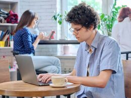 Curly guy college student typing on laptop sitting at table at coffee shop with cup of coffee.