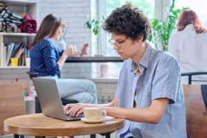 Curly guy college student typing on laptop sitting at table at coffee shop with cup of coffee.