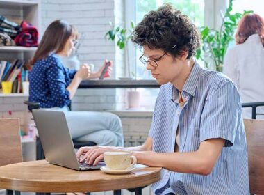 Curly guy college student typing on laptop sitting at table at coffee shop with cup of coffee.