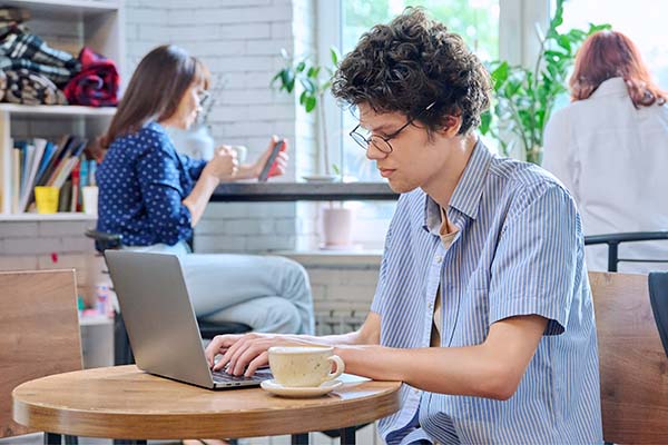 Curly guy college student typing on laptop sitting at table at coffee shop with cup of coffee.